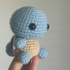 May include: A light blue crocheted turtle with black eyes. The turtle is sitting on a hand.