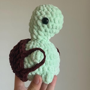 May include: A crocheted green turtle plush toy with a brown shell. The turtle has black eyes and a soft, fuzzy texture.