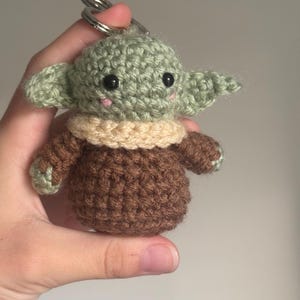 Op de afbeelding: Gehaakte sleutelhanger met een klein, groen geschilderd figuur met grote oren, zwarte ogen en een beige kraag. Het figuur heeft een bruin lichaam en is bevestigd aan een zilveren sleutelhanger. Handgemaakt item.
