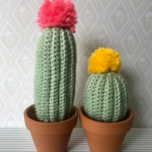 May include: Two crocheted cactus plants in terracotta pots. The larger cactus has a pink pom-pom on top and the smaller cactus has a yellow pom-pom.