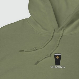 May include: Olive green hoodie with a drawstring. The hoodie features an embroidered design of a dark beer glass with a shamrock and the text "VITAMIN G" in white.