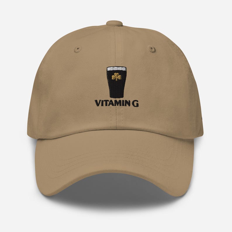 K&ouml;nnte beinhalten: Beige Baseball Cap mit gesticktem Design. Es zeigt ein schwarzes Pint Stout mit einem goldenen Kleeblatt und dem Text "VITAMIN G". Die Kappe hat einen gebogenen Schirm und einen Knopf oben.