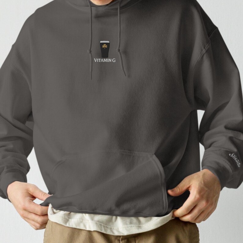 May include: A dark gray hoodie with a front pocket and a hood. The front of the hoodie features a small graphic of a pint of stout with a shamrock and the text "Vitamin G". The sleeve has the word "Sainte" embroidered.