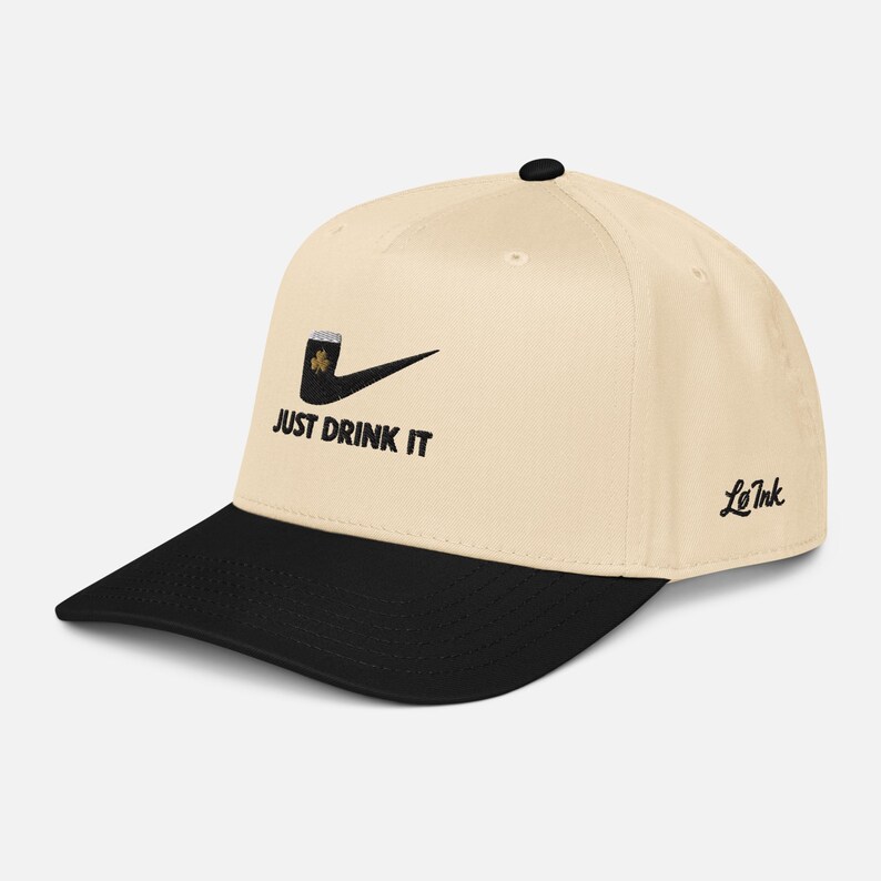 May include: Cream-colored baseball cap with a black brim. The cap features a black pipe design with a shamrock and the text "JUST DRINK IT" in black. The brand name "Le Ink" is on the side.