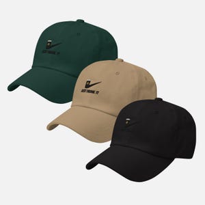 May include: Three baseball caps in dark green, tan, and black. Each cap features a beer glass logo with the text "JUST DRINK IT" in black. The caps have a curved brim and an adjustable strap.