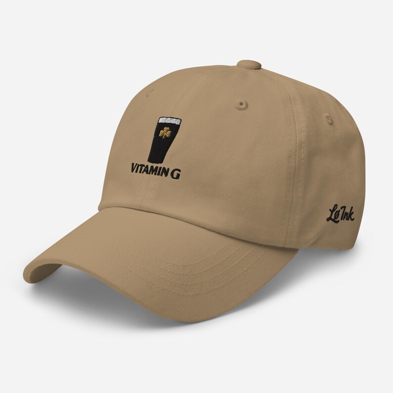 K&ouml;nnte beinhalten: Beige Baseball Cap mit gebogenem Schirm. Die Kappe hat ein gesticktes Design eines schwarzen Pintglases mit einem Kleeblatt und den Worten "VITAMIN G". Das Logo "Le Ink" ist auf der Seite gestickt.
