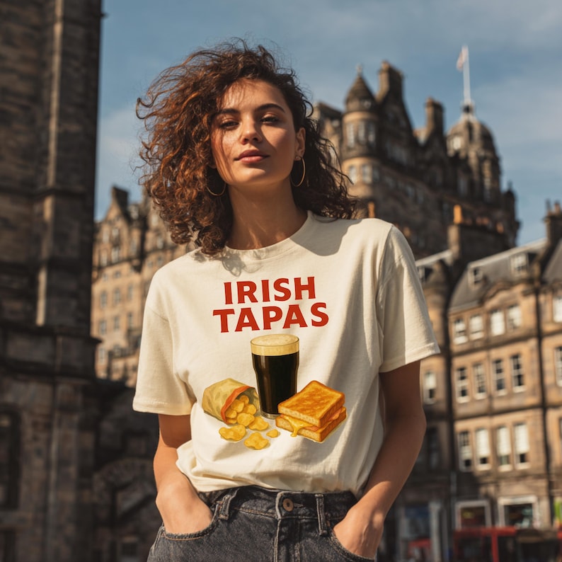 May include: Cream-colored t-shirt with the words "IRISH TAPAS" in red. The shirt features a graphic of a pint of dark beer, chips, and a grilled cheese sandwich. The person is wearing dark jeans.