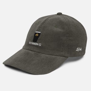 May include: Olive green corduroy baseball cap with a curved brim. The cap features an embroidered design of a pint of beer with a shamrock and the text "VITAMIN G". The brand name "Le Ink" is embroidered on the side.