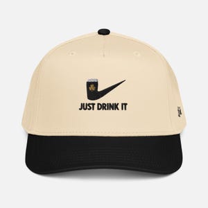 May include: A baseball cap with a cream-colored crown and a black brim. The cap features a black graphic of a beer mug with a shamrock and a checkmark, with the words "JUST DRINK IT" in black.