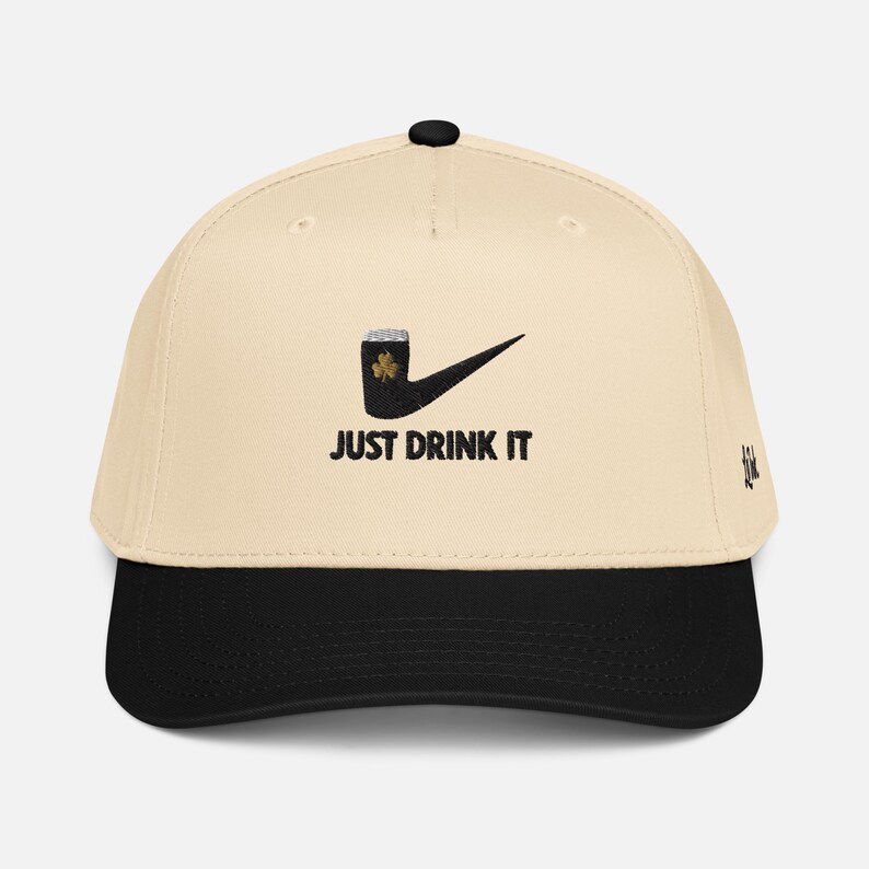 May include: A baseball cap with a cream-colored crown and a black brim. The cap features a black graphic of a beer mug with a shamrock and a checkmark, with the words "JUST DRINK IT" in black.
