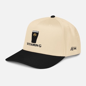 May include: A cream-colored baseball cap with a black brim. The cap features an embroidered design of a black pint glass with a gold shamrock and the text "VITAMIN G". The brand name "Le Ink" is embroidered on the side.