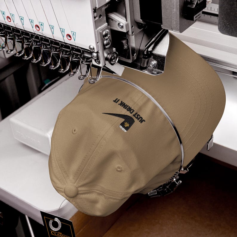 May include: A tan baseball cap with the text "JUST DRINK IT" and a logo is held in an embroidery machine. The cap is secured in a metal frame, ready for the embroidery process. The machine has multiple needles.