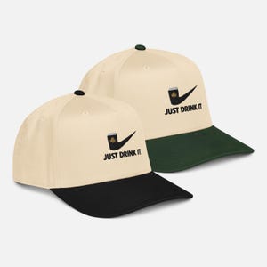 May include: Two baseball caps with a cream-colored crown and a black brim. Each cap features a black pipe design with a shamrock and the text "JUST DRINK IT". One cap has a green brim.