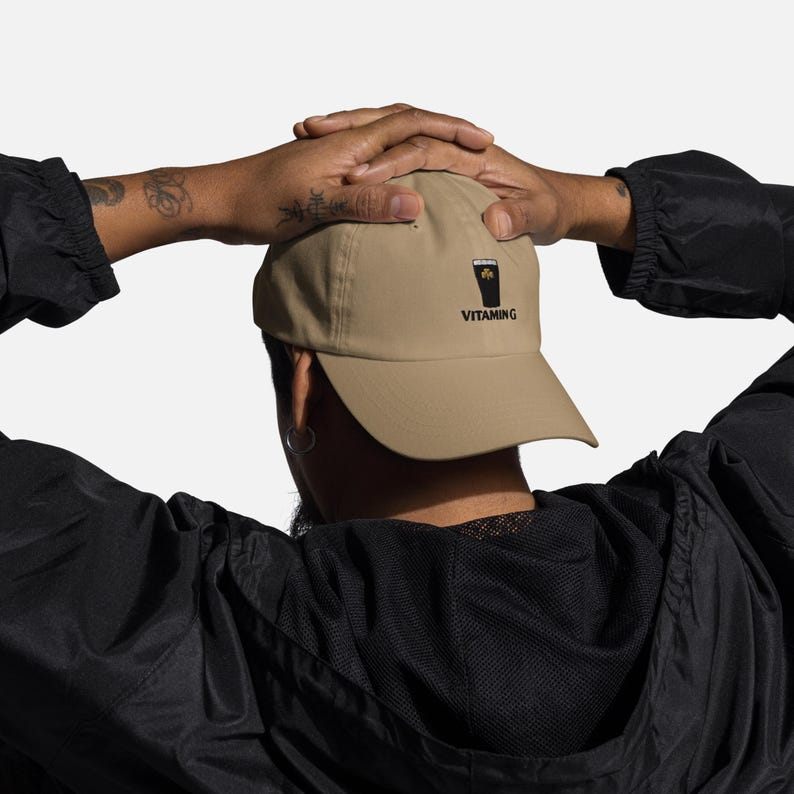 K&ouml;nnte beinhalten: Beige Baseball Cap mit einer schwarzen Grafik eines Bierkrugs und einem Kleeblatt. Das Wort "VITAMING" ist darunter in Schwarz gedruckt. Die Kappe wird von einer Person in einer schwarzen Jacke getragen.