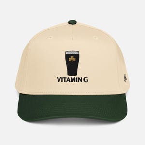 May include: Cream-colored baseball cap with a dark green brim and a black top button. The cap features an embroidered image of a pint of stout with a gold shamrock and the text "VITAMIN G" in black.