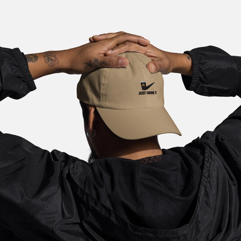May include: A tan baseball cap with a black logo and the text "JUST DRINK IT". The cap is made of a lightweight material and has a curved brim. The cap is being worn by a person.