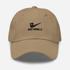 May include: Tan baseball cap with a black and green graphic of a beer mug and shamrock, with the text "JUST DRINK IT" in black. The cap has a curved brim and a classic six-panel design.