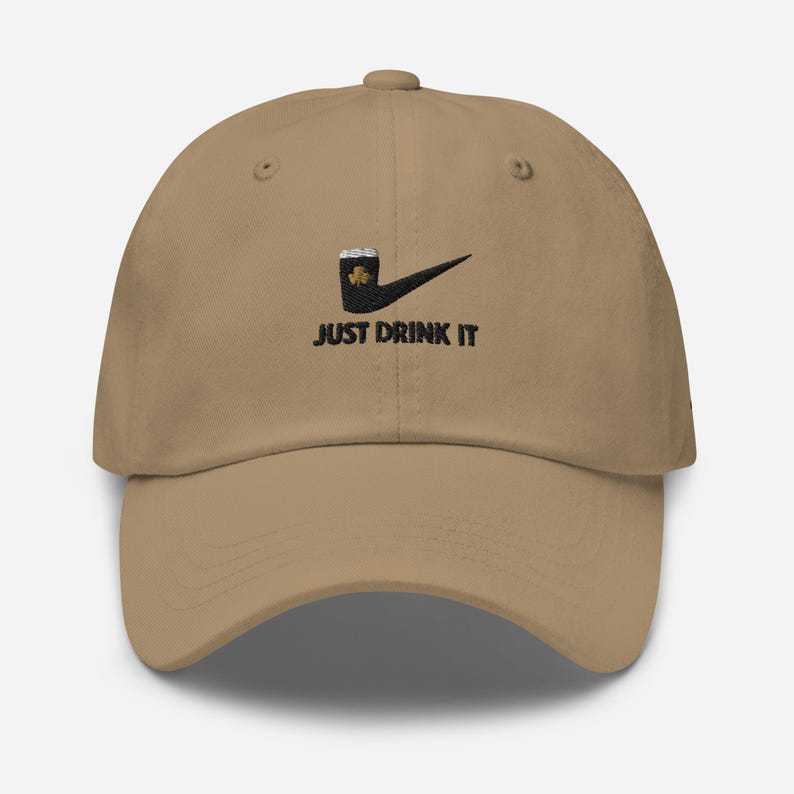 May include: Tan baseball cap with a black and green graphic of a beer mug and shamrock, with the text "JUST DRINK IT" in black. The cap has a curved brim and a classic six-panel design.