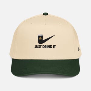 May include: Cream-colored baseball cap with a dark green brim and top button. The cap features a black graphic of a pipe with a shamrock and a checkmark, along with the text "JUST DRINK IT" in black. A small embroidered design is on the side.