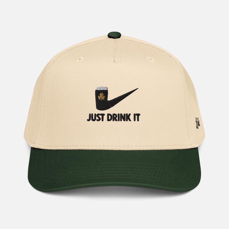 May include: Cream-colored baseball cap with a dark green brim and top button. The cap features a black graphic of a pipe with a shamrock and a checkmark, along with the text "JUST DRINK IT" in black. A small embroidered design is on the side.