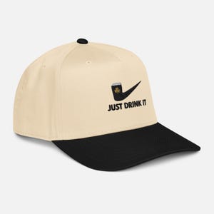 May include: A cream-colored baseball cap with a black brim. The cap features a black graphic of a pipe with a beer mug and a shamrock, with the text "JUST DRINK IT" in black.