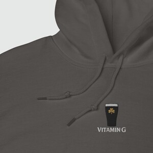 May include: A dark gray hoodie with a drawstring and a hood. The hoodie features an embroidered design of a pint of stout with a gold shamrock and the text "VITAMIN G" in white.