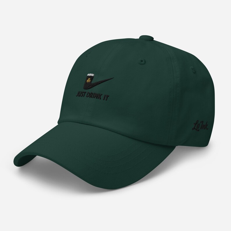May include: A dark green baseball cap with a curved brim. The cap features an embroidered design of a beer mug with a shamrock and the text "JUST DRINK IT". The brand name "LoInk" is embroidered on the side.