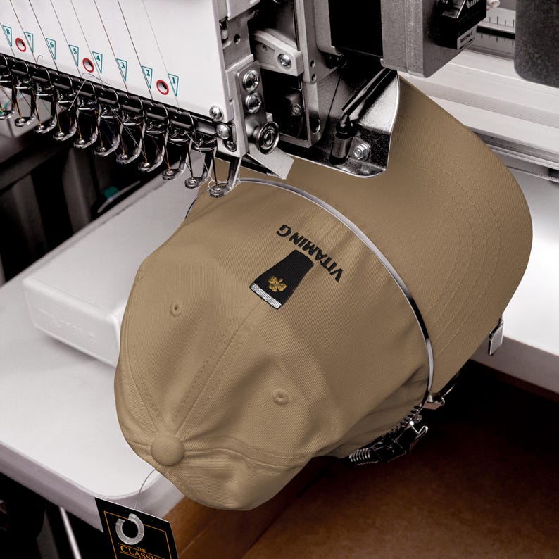 K&ouml;nnte beinhalten: Beige Baseballkappe mit dem Text "VITAMIN G" in Schwarz gestickt. Die Kappe ist in einer Stickmaschine befestigt. Die Kappe hat einen schwarzen und goldenen Aufn&auml;her. Die Maschine hat mehrere Nadeln.
