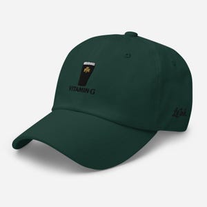 K&ouml;nnte beinhalten: Dunkelgr&uuml;ne Baseballkappe mit gebogenem Schirm. Die Kappe hat ein gesticktes Design eines schwarzen Glases mit einem goldenen Kleeblatt und dem Text "VITAMIN G". Der Markenname "Le Ink" ist auf der Seite gestickt.