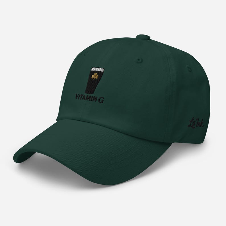 K&ouml;nnte beinhalten: Dunkelgr&uuml;ne Baseballkappe mit gebogenem Schirm. Die Kappe hat ein gesticktes Design eines schwarzen Glases mit einem goldenen Kleeblatt und dem Text "VITAMIN G". Der Markenname "Le Ink" ist auf der Seite gestickt.