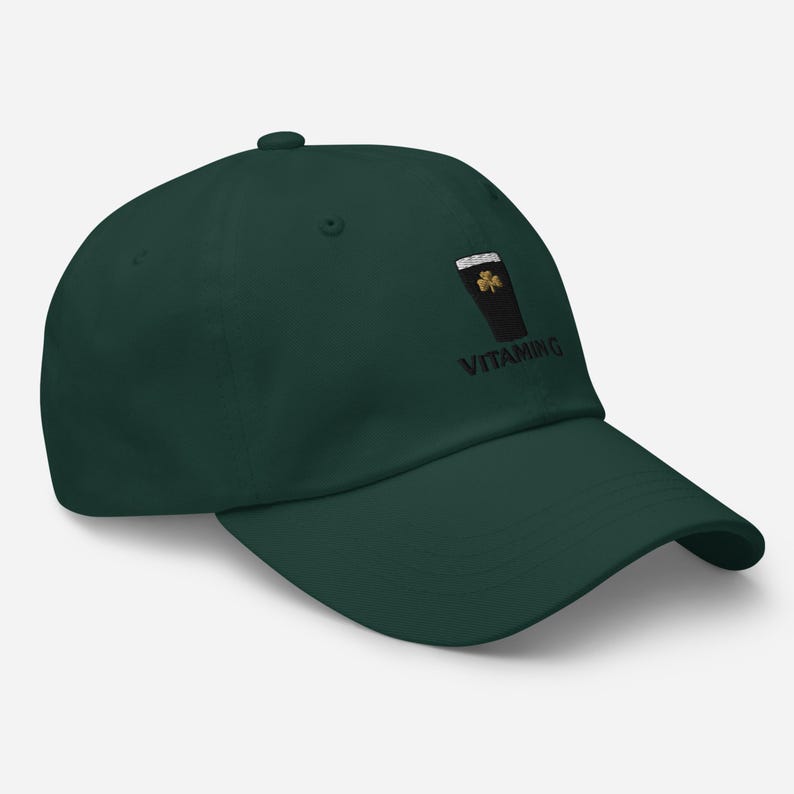K&ouml;nnte beinhalten: Dunkelgr&uuml;ne Baseballkappe mit einem gestickten Design eines Pints Stout, einem Kleeblatt und dem Wort "VITAMING" in Schwarz. Die Kappe hat einen gebogenen Schirm und einen Knopf oben.
