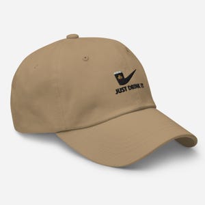 May include: A tan baseball cap with a curved brim. The cap features a black and green graphic of a pipe with a beer mug and a shamrock, along with the text "JUST DRINK IT" in black.