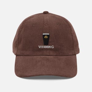 May include: Brown corduroy baseball cap with a black graphic of a pint of stout with a gold shamrock and the text "VITAMIN G" embroidered on the front. The cap has a curved brim and a button on top.