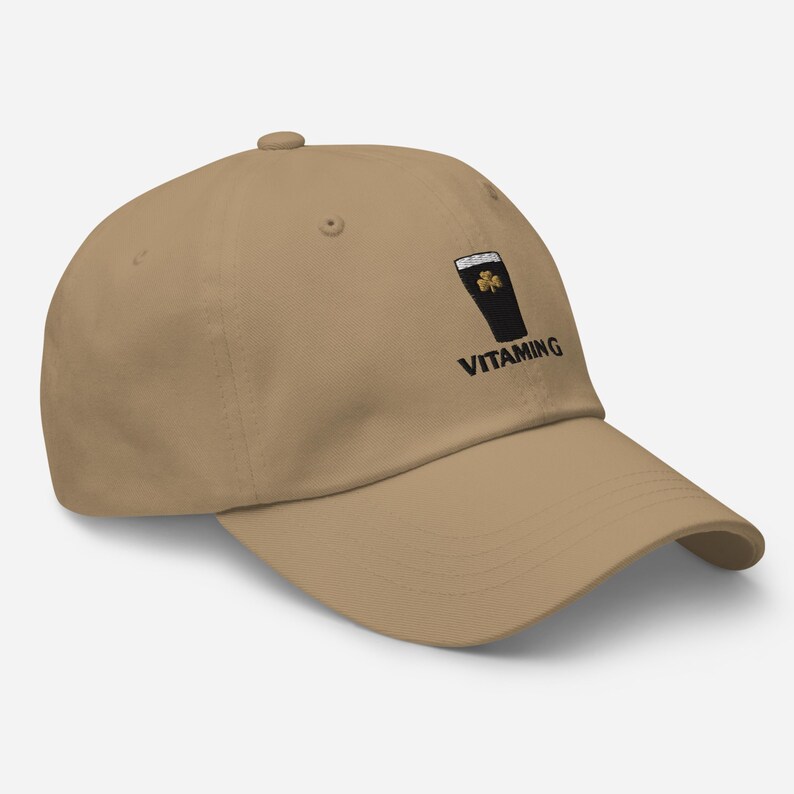 K&ouml;nnte beinhalten: Beige Baseball Cap mit gebogenem Schirm. Die Kappe zeigt eine schwarze Grafik eines Pints Stout mit einem Kleeblatt und dem Wort "VITAMING" in schwarzer Schrift.