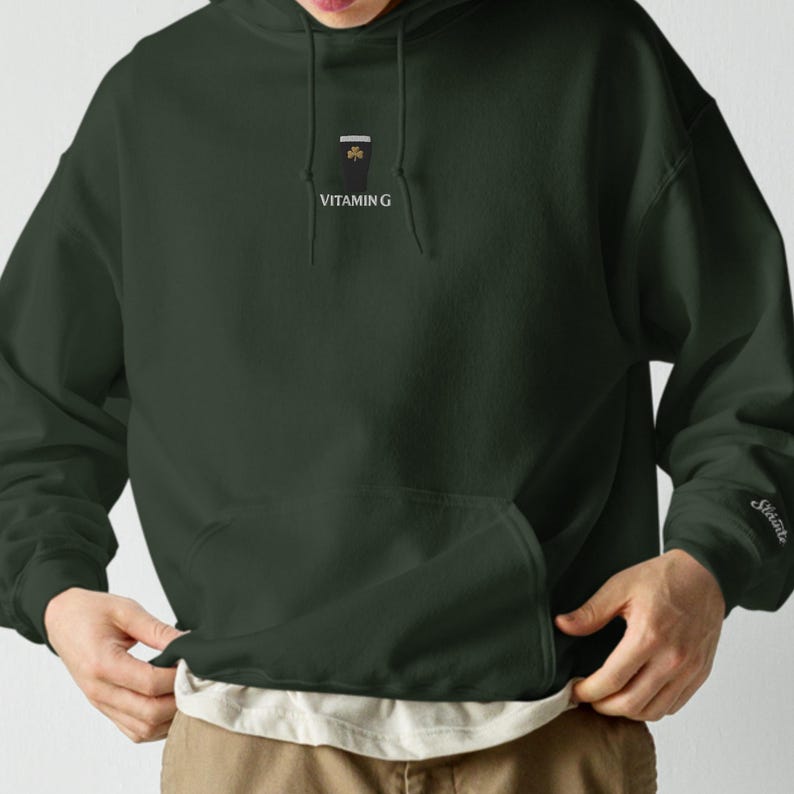May include: Dark green hoodie with a front pocket and a small graphic on the chest. The graphic includes a stylized design and the text "VITAMIN G". The sleeve has the word "Sk&ouml;nte" embroidered on it.