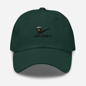 May include: A dark green baseball cap with a black embroidered design. The design features a beer mug with a shamrock and the words "JUST DRINK IT" in black. The cap has a curved brim and a button on top.