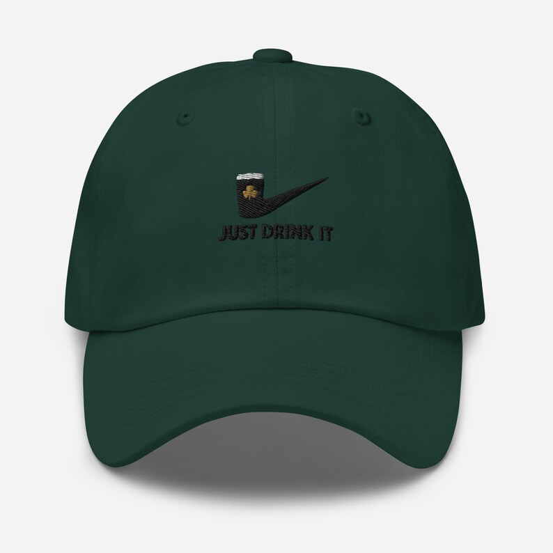 May include: A dark green baseball cap with a black embroidered design. The design features a beer mug with a shamrock and the words "JUST DRINK IT" in black. The cap has a curved brim and a button on top.