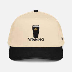 May include: A baseball cap with a cream-colored crown and a black brim. The cap features an embroidered design of a dark pint glass with a gold shamrock and the text "VITAMIN G" in black.