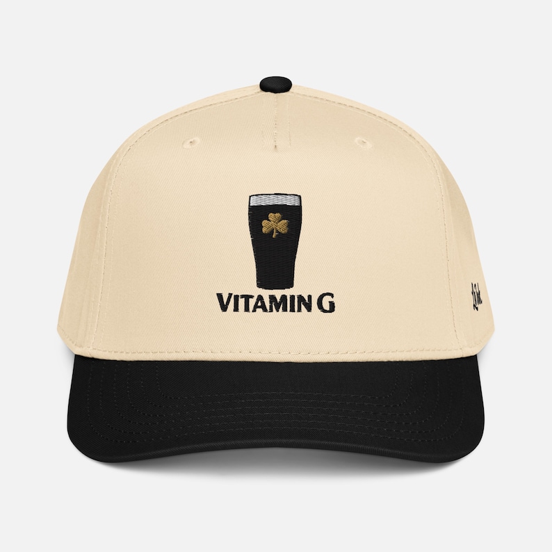 May include: A baseball cap with a cream-colored crown and a black brim. The cap features an embroidered design of a dark pint glass with a gold shamrock and the text "VITAMIN G" in black.