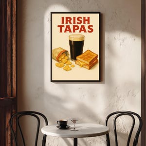 May include: A framed print featuring the words "IRISH TAPAS" in red. The image depicts a pint of dark beer, a bag of chips, and a grilled cheese sandwich. The print is displayed on a textured white wall.