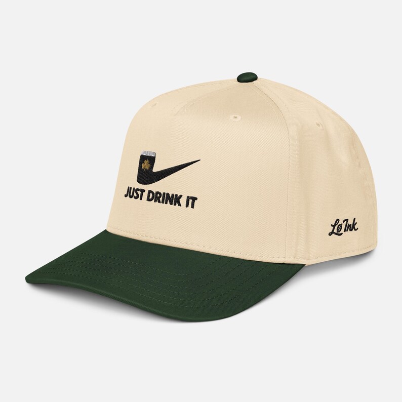 May include: Cream-colored baseball cap with a dark green brim and top button. The cap features a black graphic of a pipe with a shamrock and the text "JUST DRINK IT". The brand name "Le Ink" is on the side.