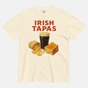 May include: Cream-colored t-shirt with the text "IRISH TAPAS" in red. The graphic features a pint of dark beer, a bag of chips, and a grilled cheese sandwich. The shirt is made of a soft, comfortable material.