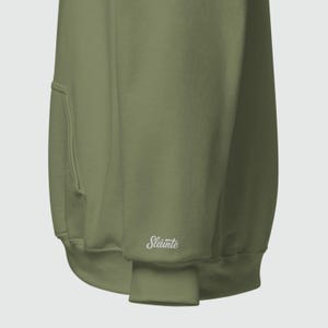 May include: Olive green hoodie with a kangaroo pocket and ribbed cuffs. The word "Sl&aacute;inte" is embroidered in white on the cuff. The hoodie is made of a soft, comfortable material.