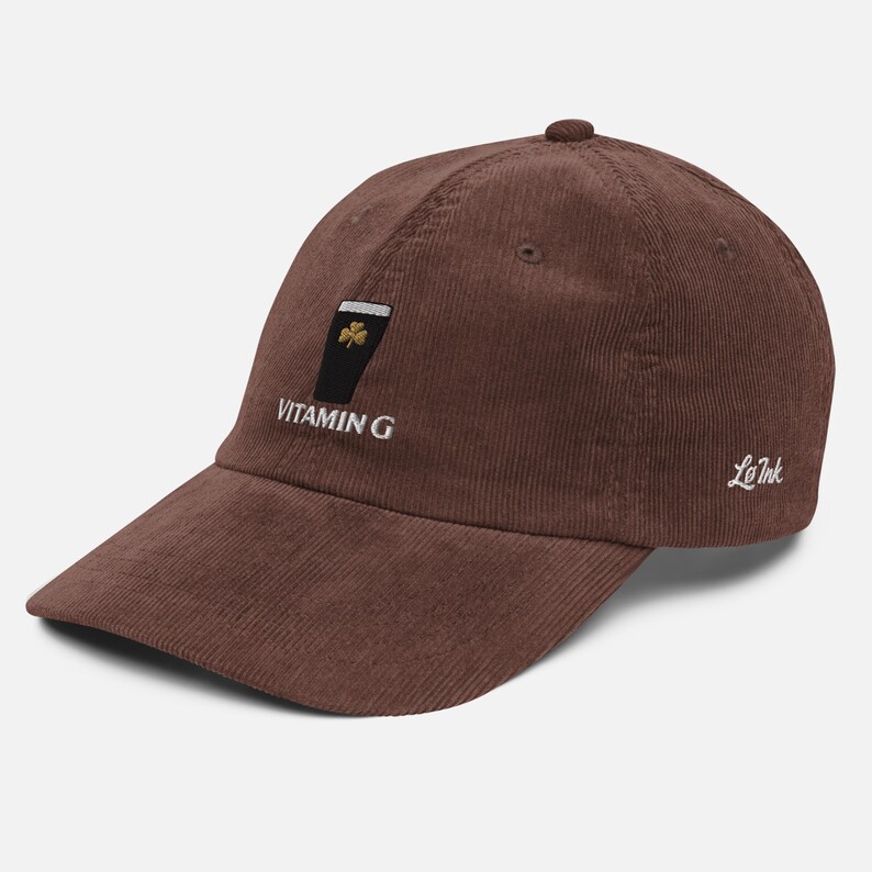 May include: Brown corduroy baseball cap with a curved brim. The cap features an embroidered design of a black pint glass with a shamrock and the text "VITAMIN G". The brand name "Le Ink" is embroidered on the side.