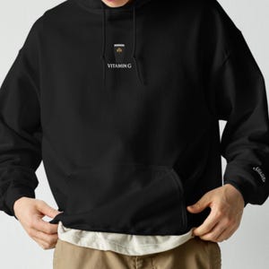 May include: Black hooded sweatshirt with a front pocket and a white drawstring. The sweatshirt features a small graphic and the text "VITAMIN G" on the chest. The sleeve has a small embroidered design.