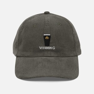 May include: Olive green corduroy baseball cap with a black pint of stout graphic and a gold shamrock. The text "VITAMIN G" is embroidered below the graphic. The cap has a curved brim and a fabric strap closure.