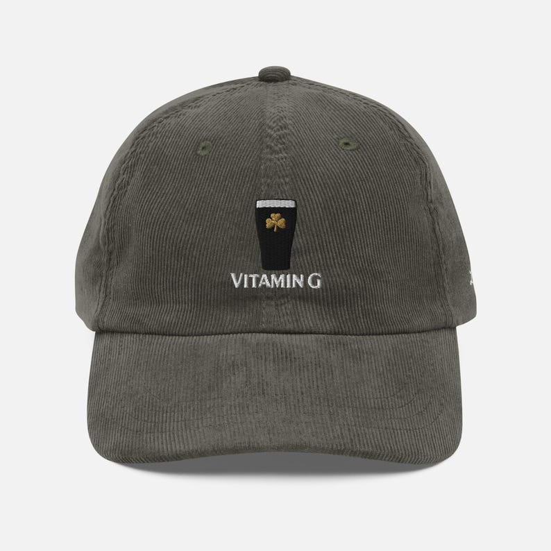 May include: Olive green corduroy baseball cap with a black pint of stout graphic and a gold shamrock. The text "VITAMIN G" is embroidered below the graphic. The cap has a curved brim and a fabric strap closure.