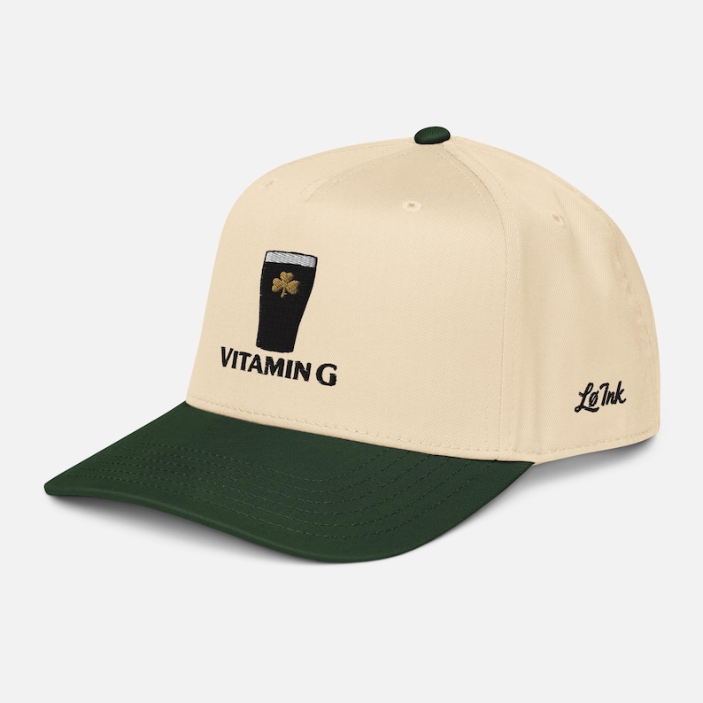 May include: Cream-colored baseball cap with a dark green brim and top button. The cap features an embroidered design of a black pint glass with a shamrock and the text "VITAMIN G". The brand name "Le Ink" is on the side.