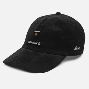 May include: Black corduroy baseball cap with a curved brim. The cap features the embroidered text "VITAMIN G" in white, along with a small gold shamrock and the "Le Ink" logo. The cap is a stylish accessory.