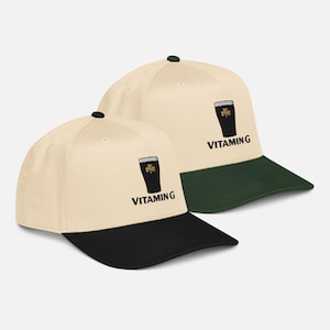 May include: Two baseball caps with a cream-colored crown and a black brim. Each cap features an embroidered black pint of stout with a shamrock and the word "VITAMING" in black letters. One cap has a green brim.
